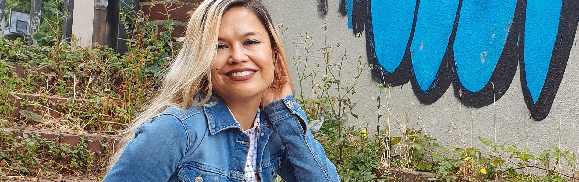 Eine Frau sitzt auf einer Treppe. Sie sieht freundlich aus und trägt eine Jeansjacke. Im Hintergrund sieht man ein blaues Graffiti.