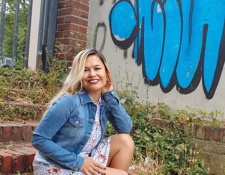 Eine Frau sitzt auf einer Treppe. Sie sieht freundlich aus und trägt eine Jeansjacke. Im Hintergrund sieht man ein blaues Graffiti.