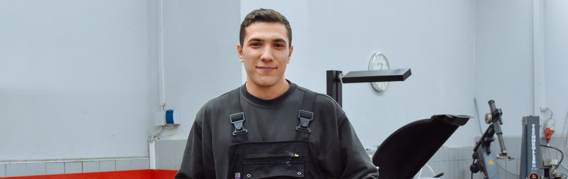 Ein junger man steht in einer Werkstatt. Er trägt Arbeitskleidung und sieht glücklich aus.