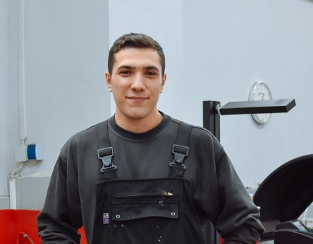 Ein junger man steht in einer Werkstatt. Er trägt Arbeitskleidung und sieht glücklich aus.