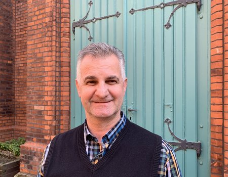 Ein freundlicher älterer Mann lächelt in die Kamera. Er steht vor einer grünen Kirchentür.