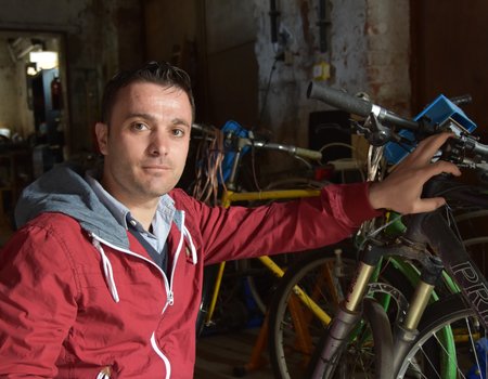 Ein junger Mann mit roter Jacke kniet for einem Fahrrad.