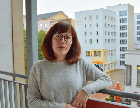 Eine junge Frau steht auf ihrem Balkon. Sie schaut ernst und es ist schlechtes Wetter.