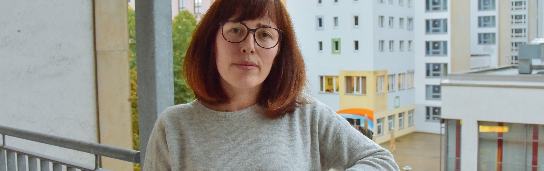 Eine junge Frau steht auf ihrem Balkon. Sie schaut ernst und es ist schlechtes Wetter.
