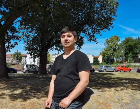 Ein freundlicher Mann steht an einen Stein gelehnt. Er trägt ein schwarzes T-Shirt und eine blaue Jeans.