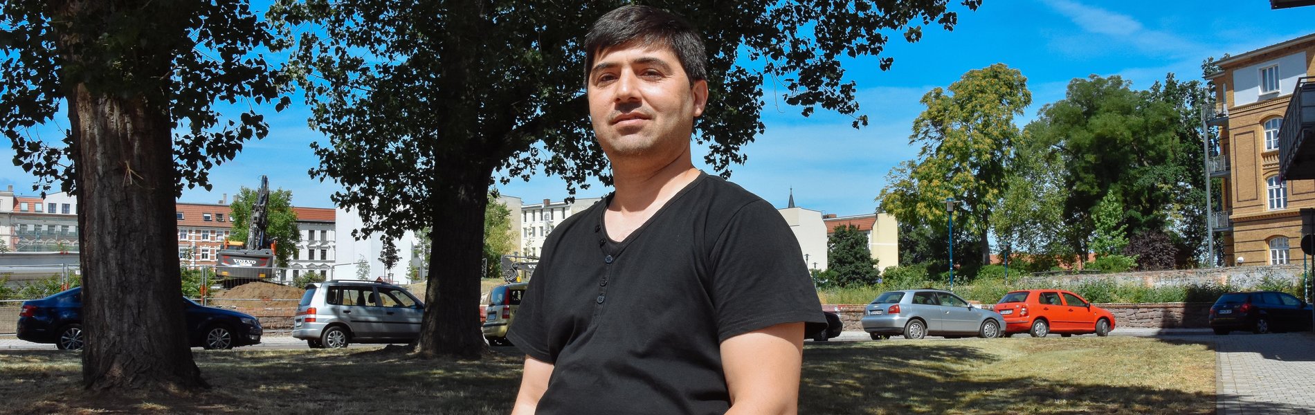Ein freundlicher Mann steht an einen Stein gelehnt. Er trägt ein schwarzes T-Shirt und eine blaue Jeans.
