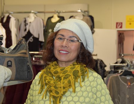 Eine ältere Dame steht in ihrem Modegeschäft und trägt einen großen Hut.