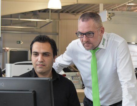 Im Bildvordergrund sind zwei Personen an einem Schreibtisch zusehen. Ein jüngerer Mann sitze an einem Computer. Er ist der Auszubildende. Hinter ihm steht der Ausbilder des Autohauses und zeigt einem Arbeitsschritt. Beide sind konzentriert. Der junge Mann trägt ein schwarzes Hemd und der ältere ein weißes. Außerdem trägt der Ausbilder eine Brille und hat einen grünen Schlips des Autohauses um. Im Hintergrund stehen Autos der Firma Skoda.