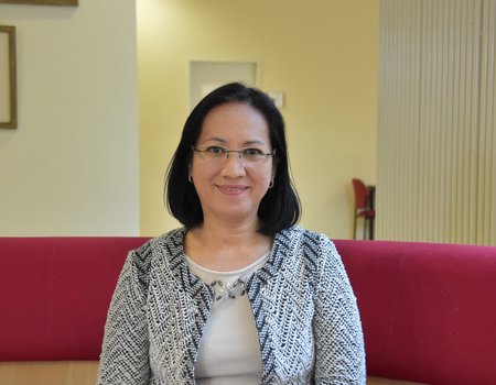Eine asiatische Frau sitzt auf einem Sofa. Sie trägt  eine Brille und eine karierte Weste.