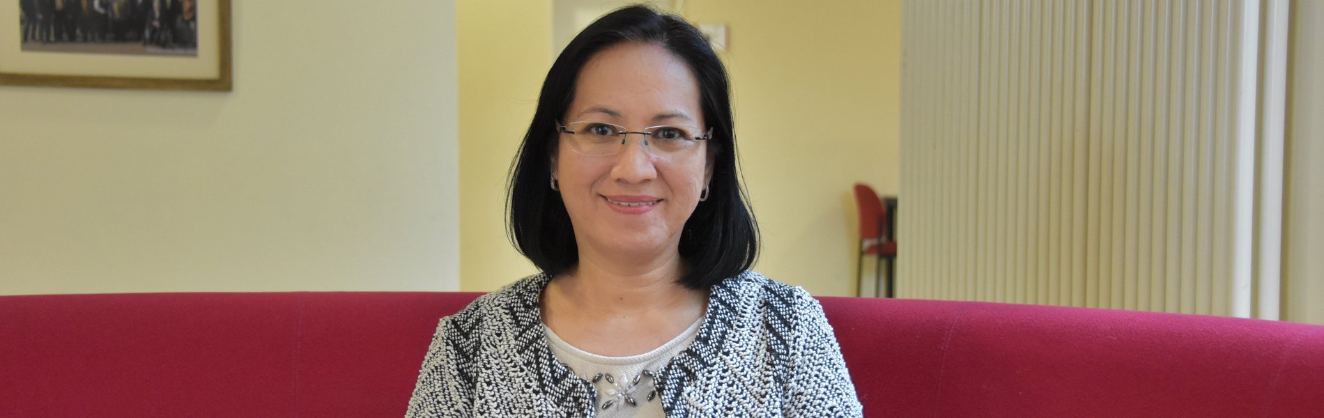 Eine asiatische Frau sitzt auf einem Sofa. Sie trägt  eine Brille und eine karierte Weste.