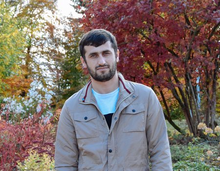 Ein junger Mann mit Vollbart steht in einem Park. Die Bäume haben bunte Blätter.