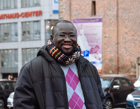 Ein junger schwarzer Mann steht auf einem Markplatz. Er trägt dicke Wintersachen.