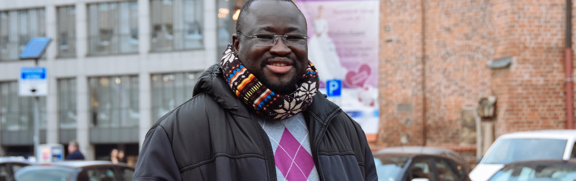 Ein junger schwarzer Mann steht auf einem Markplatz. Er trägt dicke Wintersachen.