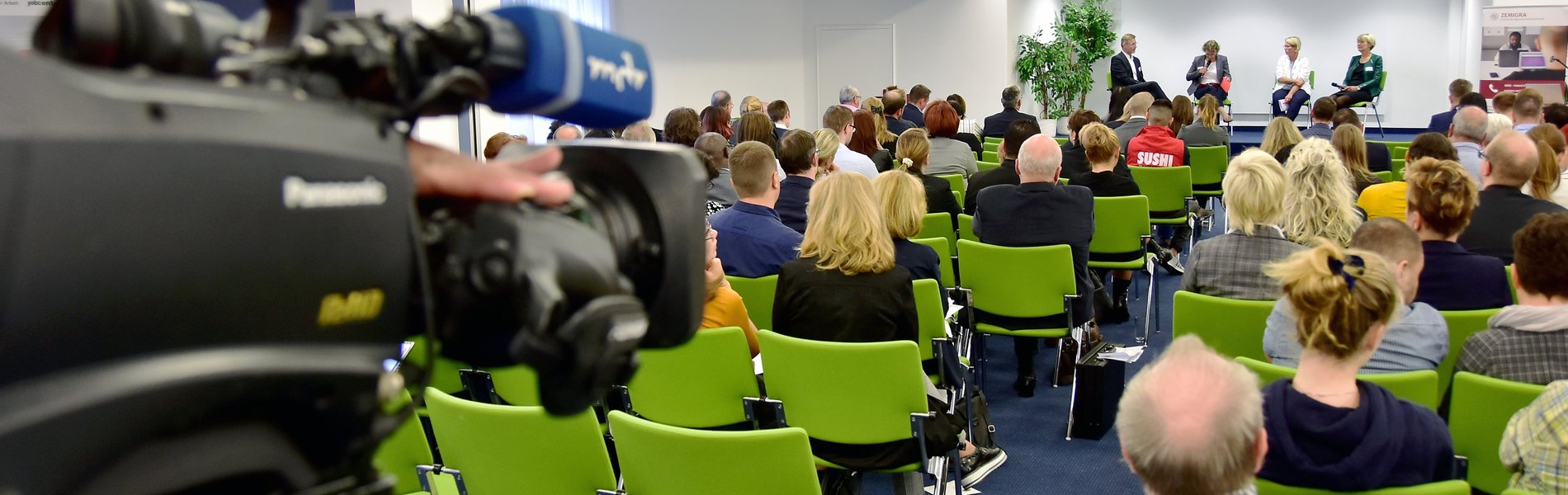 Das Foto zeigt eine Plenumssituation. Aus dem Blickwinkel einer TV-Kamera zeigt es einen Raum mit Stuhlreihen. Auf diese haben Zuhörerinnen und Zuhörer Platz genommen. Im hinteren Bereich des Raumes findet gerade ein Podiumsgespräch statt. Moderiert von Wiebke Reyels besprechen Staatssekretärin Susi Möbbeck, Landesministerium für Arbeit, Soziales und Integration, Ines Rudolph, Fachdienstleiterin der Ausländerbehörde Magdeburg, sowie Krzysztof Blau, Geschäftsführer der Auslandsgesellschaft Sachsen-Anhalt e. V., Möglichkeiten und Grenzen des neuen Fachkräfteeinwanderungsgesetzes.