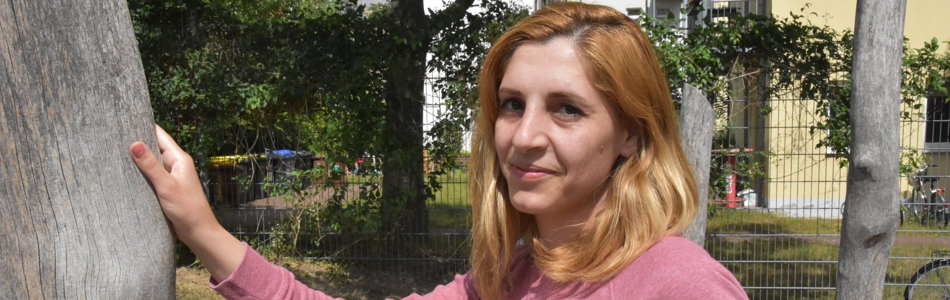 Das Foto zeigt eine Frau. Ihr schulterlanges dunkelblondes Haar umrahmt ihr Gesicht. Sie trägt einen lachsfarbenen Pullover. Mit passendem Nagellack und passendem Lippenstift. Sie lehnt an einem Baum. Die Sonne scheint. Sie schaut erwartungsvoll in die Kamera. 