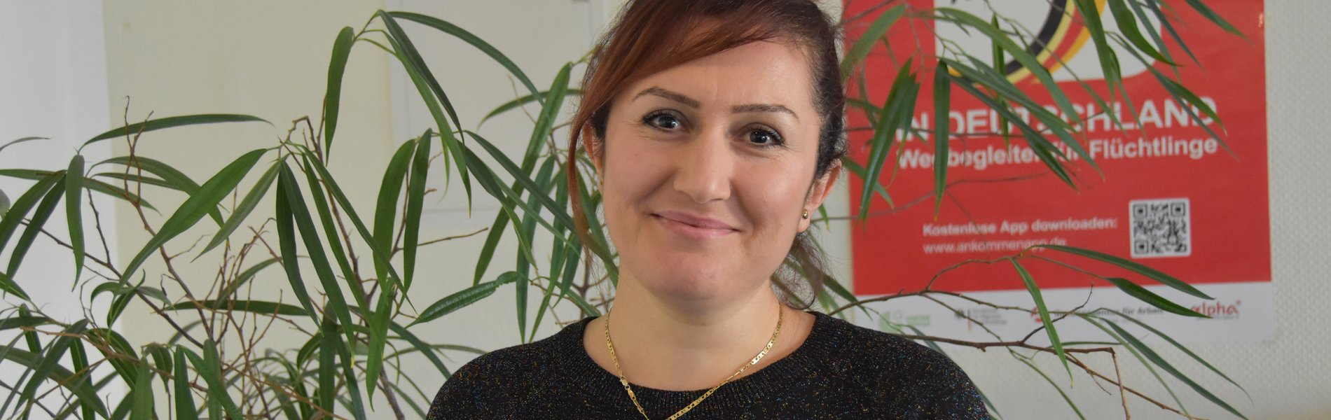 Eine junge Frau steht vor einer Pflanze und lächelt in die Kamera. Sie hat braune Haare und trägt einen schwarzen Pullover. Um den Hals hat sie eine goldene Kette. Sie macht einen glücklichen Eindruck.