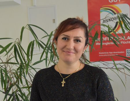 Eine junge Frau steht vor einer Pflanze und lächelt in die Kamera. Sie hat braune Haare und trägt einen schwarzen Pullover. Um den Hals hat sie eine goldene Kette. Sie macht einen glücklichen Eindruck.