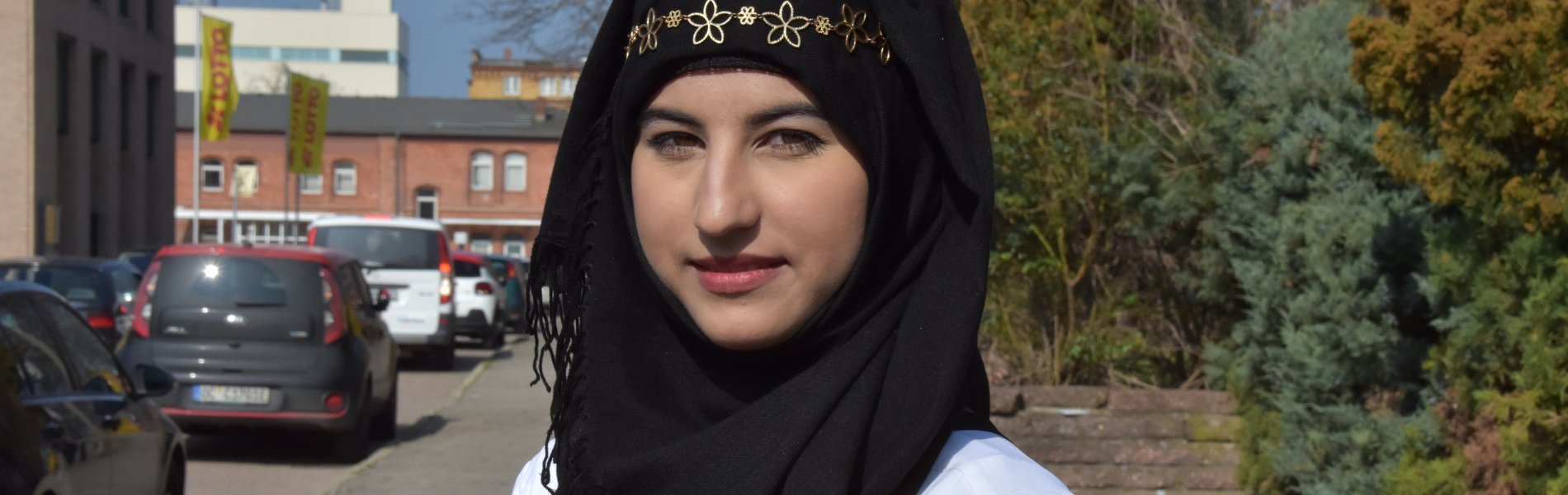 Ein junges Mädchen steht auf der Straße. Sie trägt ein schwarzes Kopftuch mit goldenem Schmuck.