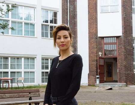 Eine junge Frau steht in einem Innenhof. Sie ist schwarz gekleidet und stark geschminkt.