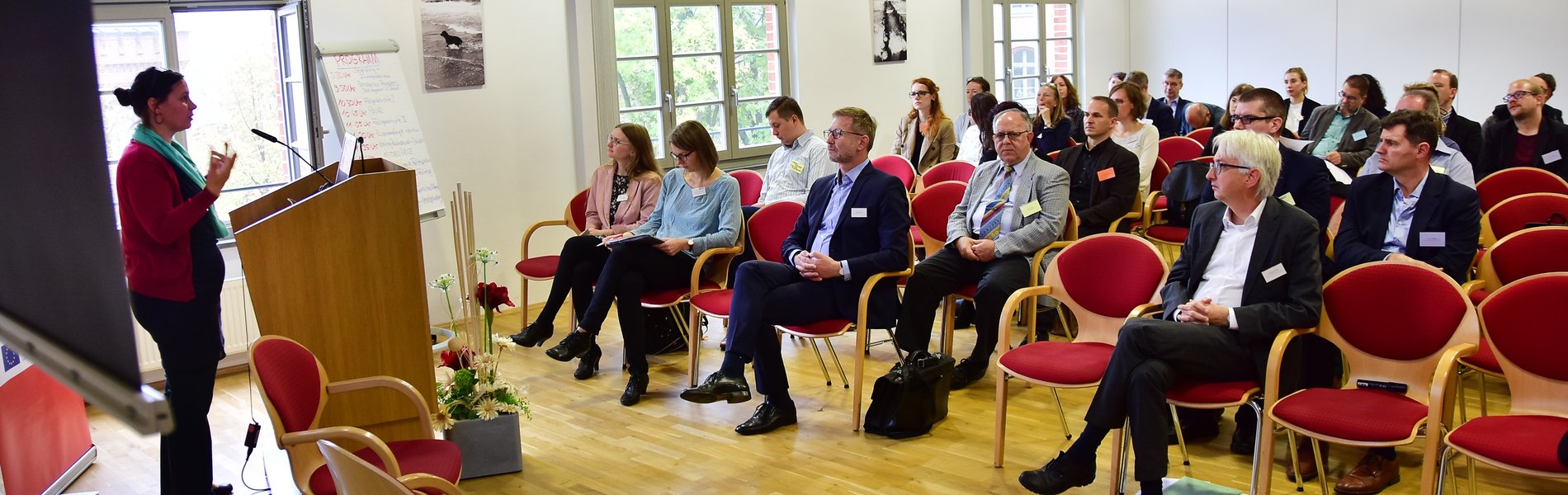 Das Foto zeigt einen Veranstaltungsraum im Ministerium für Arbeit, Soziales und Integration Sachsen-Anhalt. Dr. Franka Kretschmer begrüßt die Teilnehmenden des Fachtags an einem Rednerpult stehend. In den gut gefüllten Reihen des Fachpublikums sind u. a. Wolfgang Beck, Abteilungsleiter Arbeit und Integration des Ministeriums für Arbeit, Soziales und Integration Sachsen-Anhalt, Krzysztof Blau, Geschäftsführer der Auslandgesellschaft Sachsen-Anhalt e.V. (AGSA), Pauline Lendrich (Faire Integration) und Anne Hafenstein (BemA) zu erkennen.
