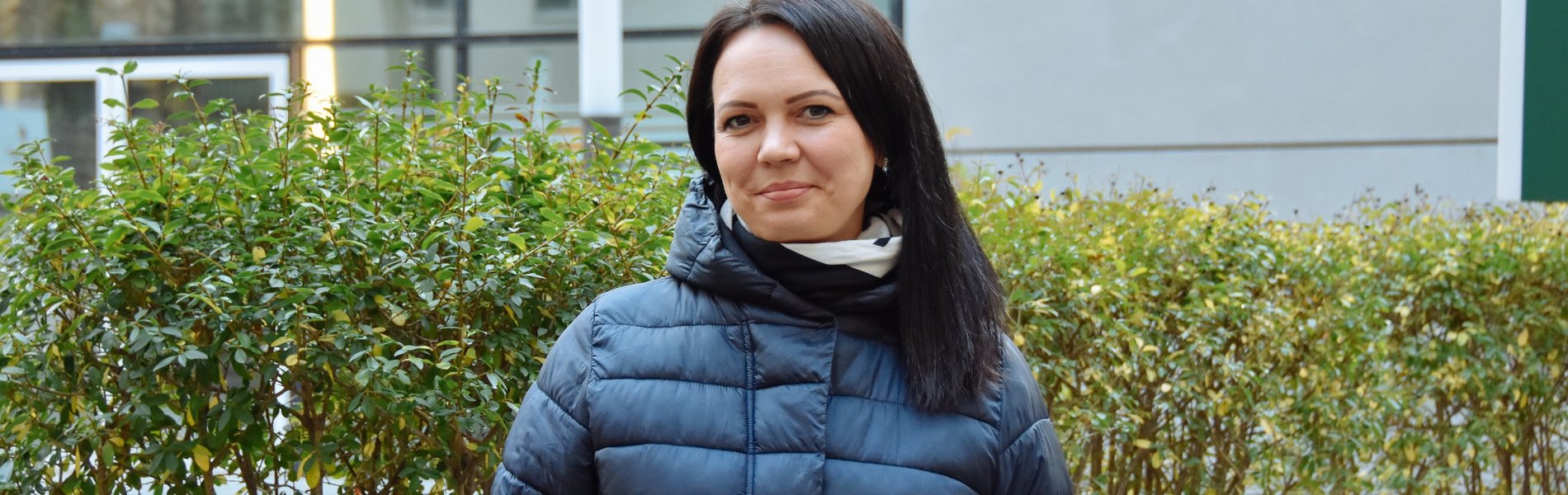 Ein junge Frau steht in Winterjacke vor einem Busch. Sie hat schwarze Haare und lächelt.