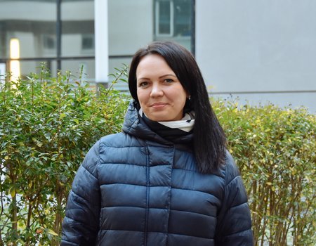 Ein junge Frau steht in Winterjacke vor einem Busch. Sie hat schwarze Haare und lächelt.