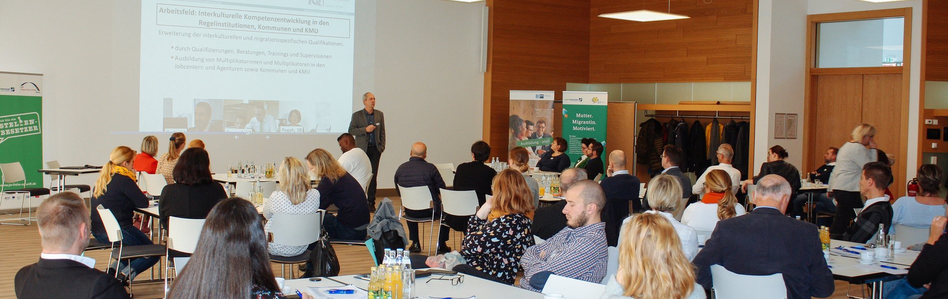 Das Foto zeigt einen Veranstaltungsraum. Ronald Schütz, Projektleiter des Angebotes Servicestelle IQ "Interkulturelle Beratung und Trainings" der Jugendwerkstatt "Frohe Zukunft" Halle-Saalekreis e.V., steht vor gut gefüllten Tischen mit Unternehmensvertreterinnen und Unternehmensvertretern. Er stellt anhand einer Präsentation die Angebote des IQ Netzwerks vor.