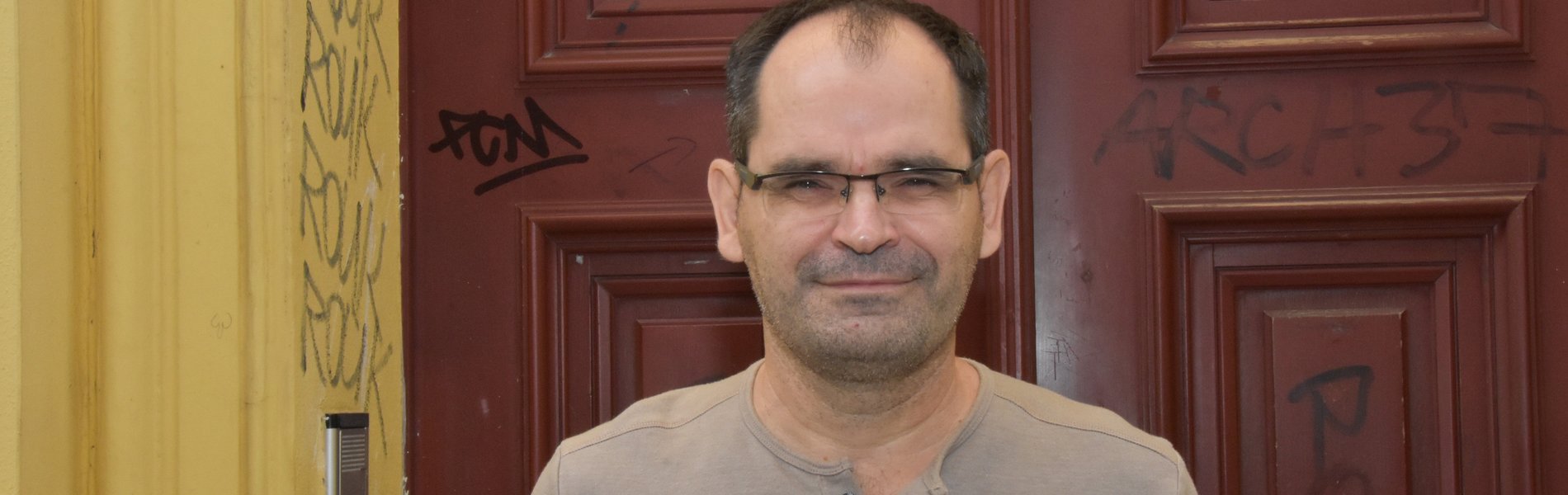 Ein 52 jähriger Mann schaut frontal in die Kamera. Das Foto zeigt den Mann nur bis zu Brust. Er hat braune Haare, eine Brille und ein braunes Shirt an. Die Sonne scheint ihn etwas zu blenden. Im Hintergrund erkennt man, eine braune Holztür. Er steht in einem Hauseingang.