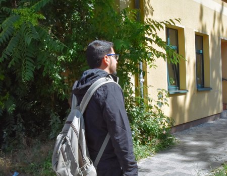 Ein junger Mann steht mit dem Rücken zur Kamera. Er hat einen Rucksack auf dem Rücken und trägt eine Brille.