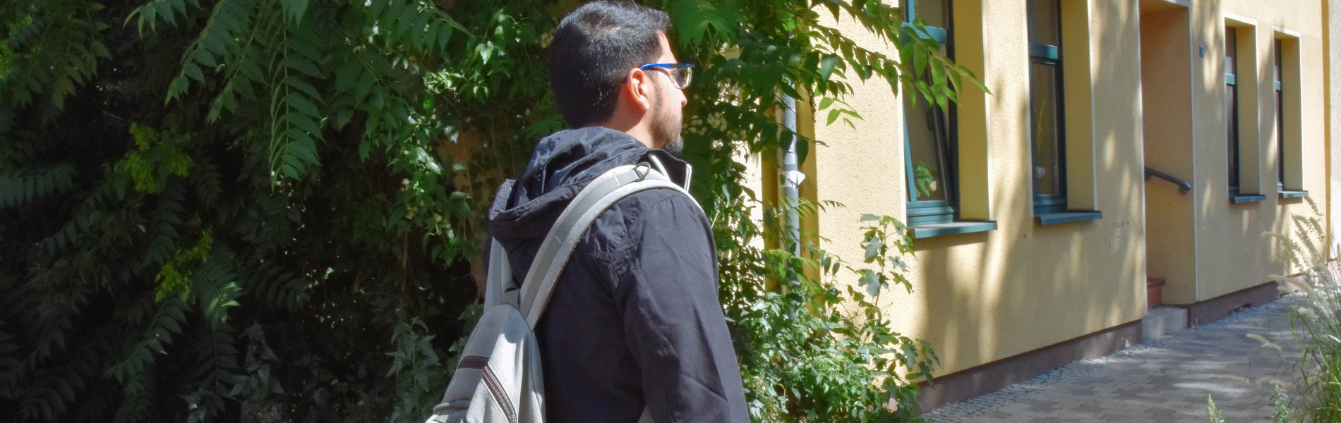 Ein junger Mann steht mit dem Rücken zur Kamera. Er hat einen Rucksack auf dem Rücken und trägt eine Brille.