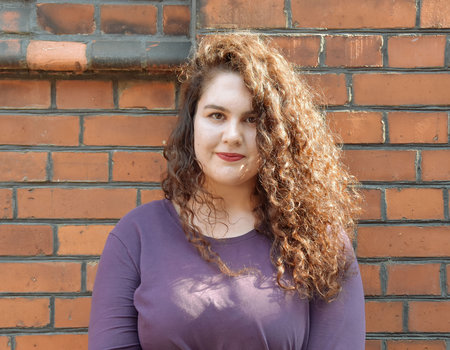 Eine junge Frau steht vor einer roten Steinwand. Sie hat langes , braunes Haar mit Locken.