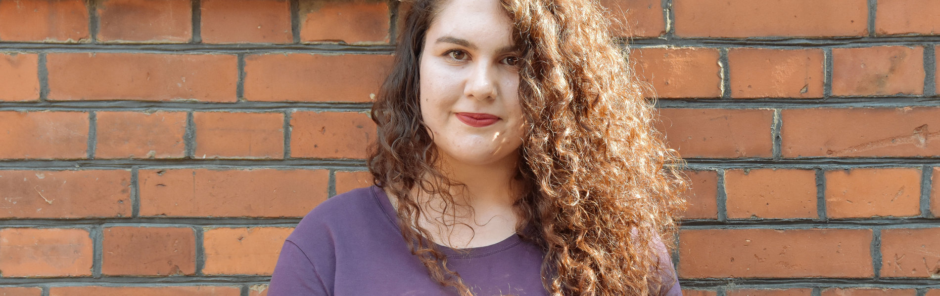 Eine junge Frau steht vor einer roten Steinwand. Sie hat langes , braunes Haar mit Locken.