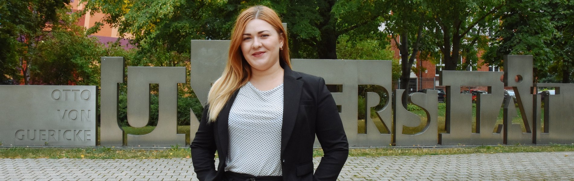 Eine junge Frau steht auf einem Platz. Sie hat blonde Haare und lächelt.