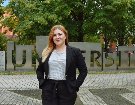 Eine junge Frau steht auf einem Platz. Sie hat blonde Haare und lächelt.